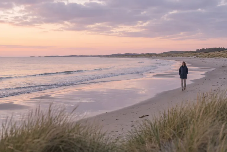 En ensam person i mörk jacka promenerar längs en öppen sandstrand på västkusten i skymningen
